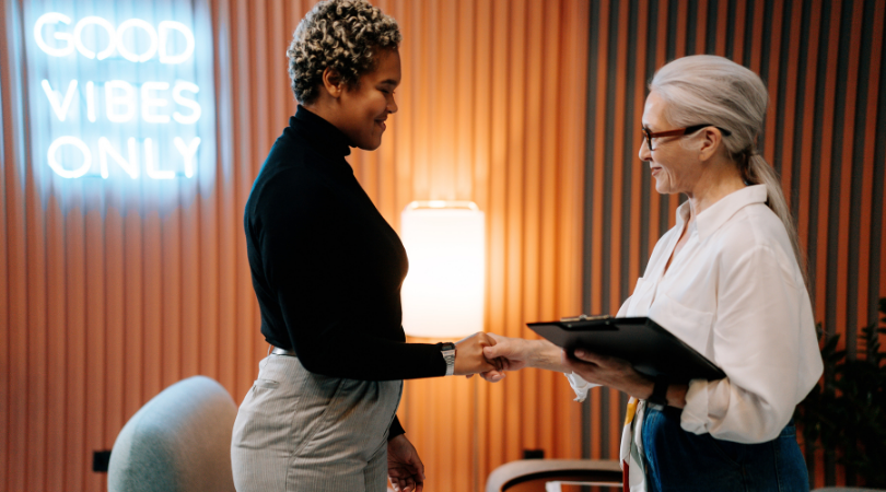 two women shake hands in a modern office, a neon light with the words 'no bad vibes' hangs above them