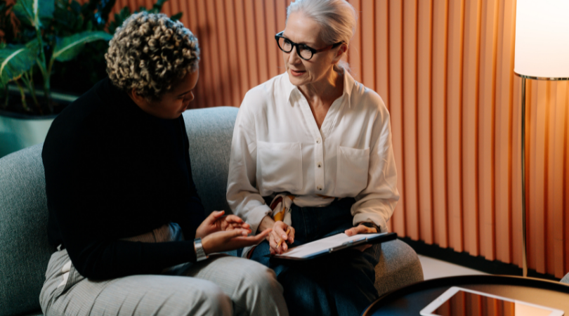 two women in office sit on a grey sofa discussing writing on a clipboard